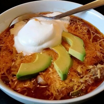 Chili served in white bowl, topped with 3 avocado slices and scoop of sour cream